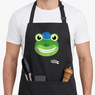 Tablier de cuisine noir avec tête de grenouille verte souriante portant une casquette bleue et le logo Gecko's Garage en bas à droite.