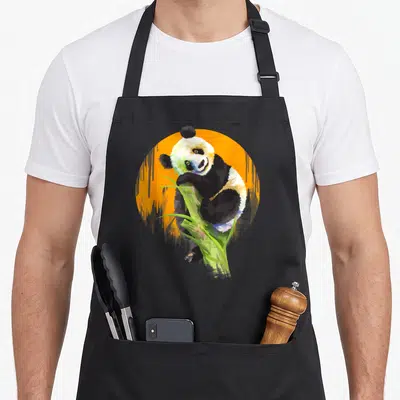 Tablier de cuisine noir avec illustration colorée d'un panda grimpant sur une tige d'herbe verte devant un disque orange.