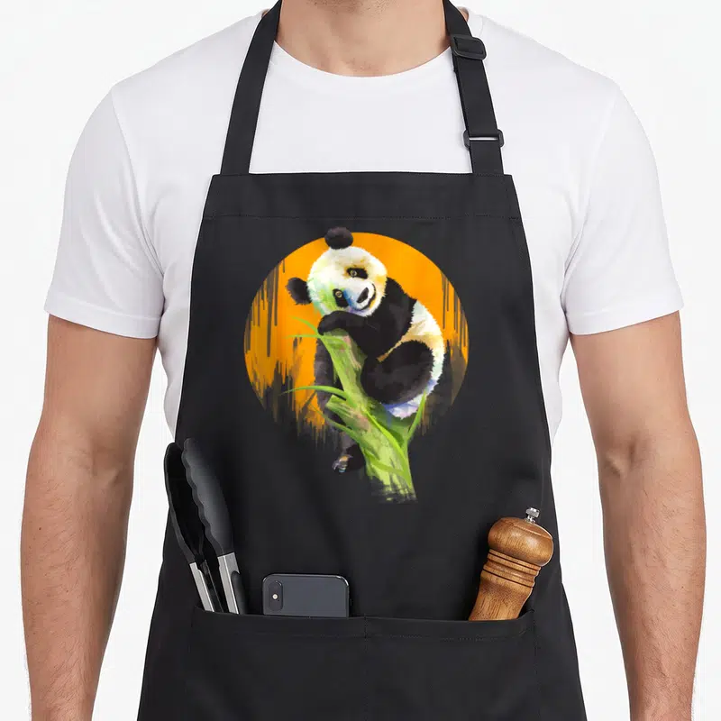 Tablier de cuisine noir avec illustration colorée d'un panda grimpant sur une tige d'herbe verte devant un disque orange.