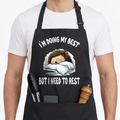 Tablier de cuisine noir avec un hérisson endormi sur un coussin blanc et du texte en anglais.