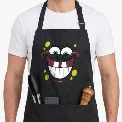 Tablier de cuisine noir avec un grand smile face souriant de Bob l'éponge aux dents visibles et bulles jaunes.