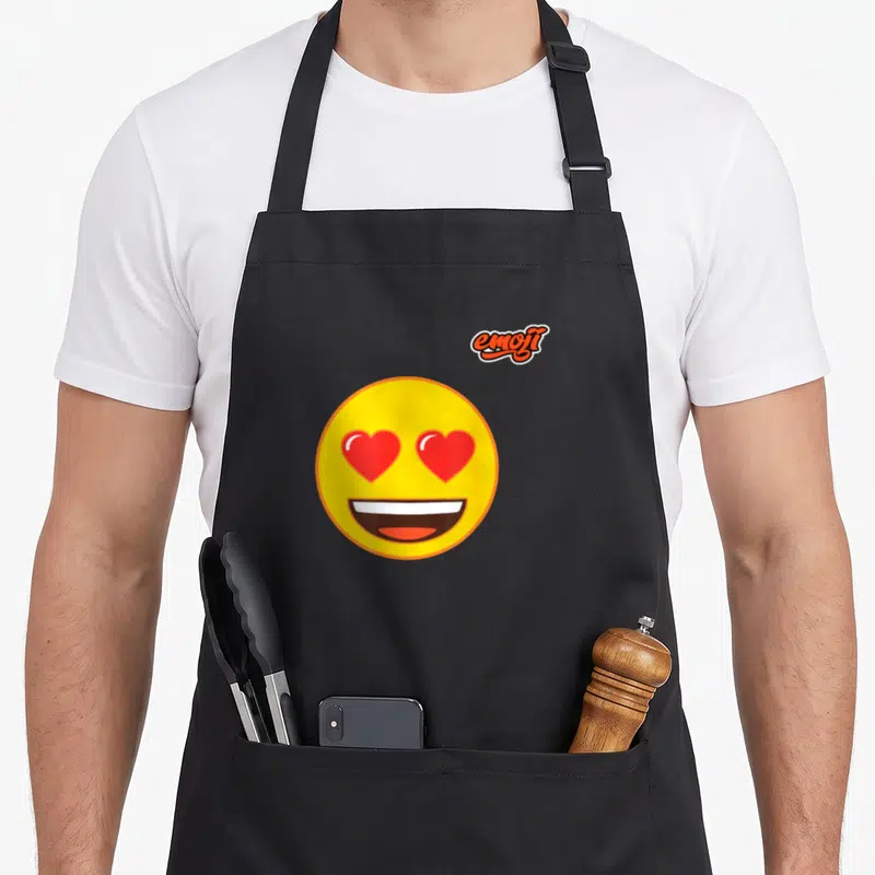 Tablier de cuisine noir avec un grand emoji jaune aux yeux en cœur rouge et une large bouche souriante.