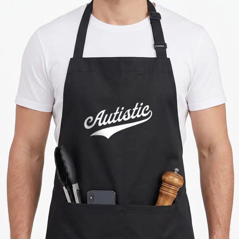 Tablier de cuisine noir avec l'inscription Autistic en lettres blanches cursives et une ligne stylisée sous le texte.