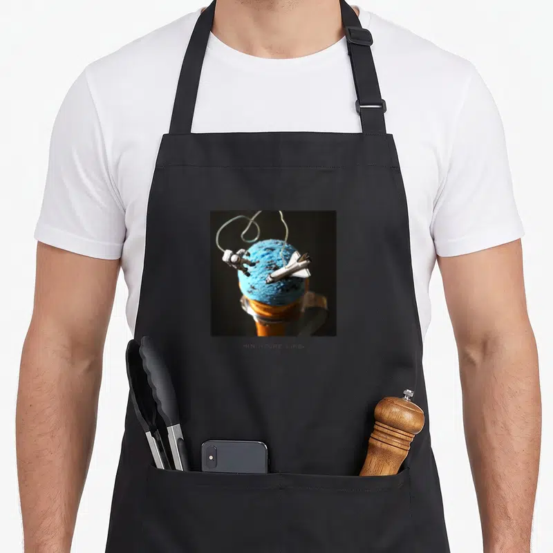 Tablier de cuisine noir avec une boule de sorbet bleue, un astronaute miniature et une fusée spatiale sur le devant.