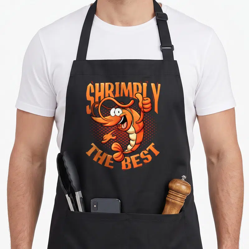 Tablier de cuisine noir avec une crevette orange cartoon faisant un signe de thumbs up et le texte Shrimply The Best.