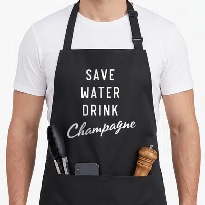 Tablier de cuisine noir avec l'inscription Save Water Drink Champagne en blanc et typographie mixte.