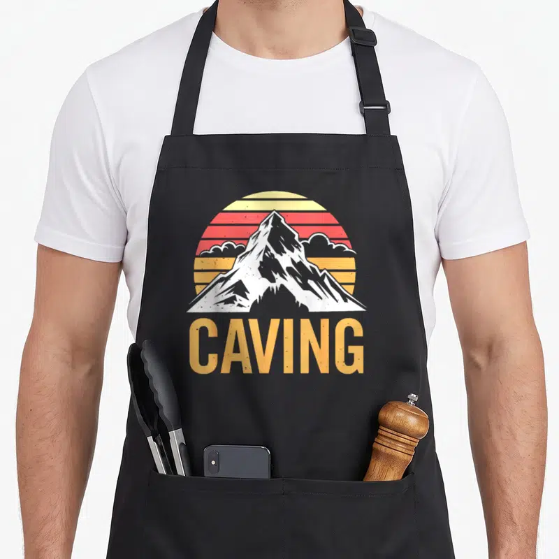 Tablier de cuisine noir avec illustration vintage d'un sommet enneigé sur fond de soleil rayé et le mot Caving en orange.