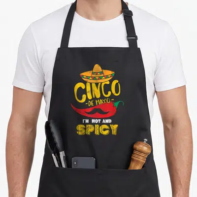 Tablier de cuisine noir avec illustration d'un piment rouge portant un sombrero et une moustache, complétée par du texte jaune et blanc.