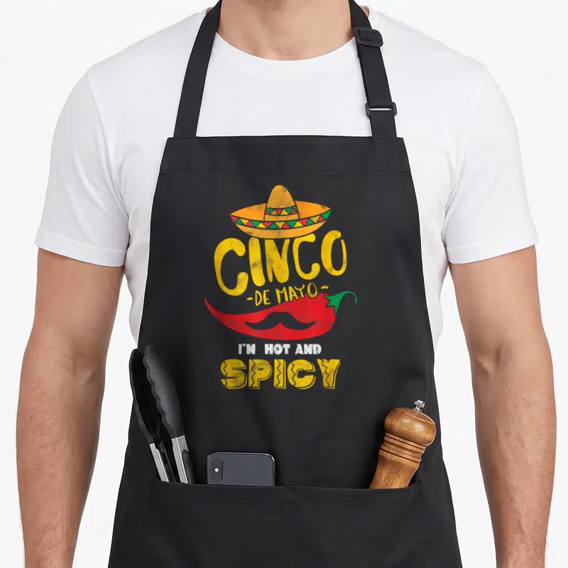 Tablier de cuisine noir avec illustration d'un piment rouge portant un sombrero et une moustache, complétée par du texte jaune et blanc.