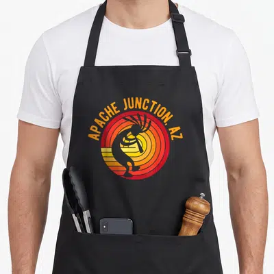 Tablier de cuisine noir avec illustration circulaire orange et rouge représentant un Kokopelli dansant au-dessus de rayons de soleil stylisés.