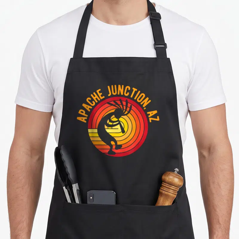 Tablier de cuisine noir avec illustration circulaire orange et rouge représentant un Kokopelli dansant au-dessus de rayons de soleil stylisés.