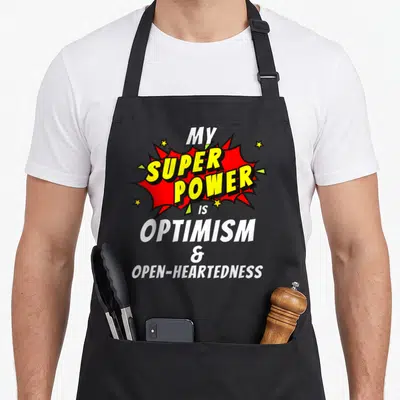 Tablier de cuisine noir avec une explosion rouge et jaune portant l'inscription My super power is optimism and open-heartedness.