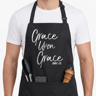 Tablier de cuisine noir avec l'inscription blanche Grace Upon Grace et la référence John 1:16 en bas à droite.