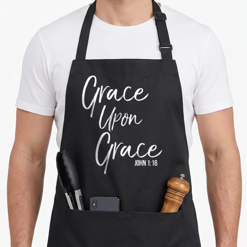 Tablier de cuisine noir avec l'inscription blanche Grace Upon Grace et la référence John 1:16 en bas à droite.