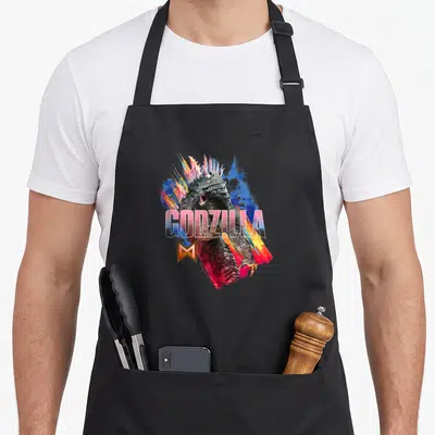 Tablier de cuisine noir avec illustration artistique de Godzilla et le logo M coloré.