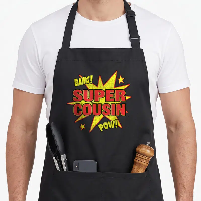 Tablier de cuisine noir avec une illustration en forme d'explosion jaune et rouge portant les mots Super Cousin Bang et Pow.