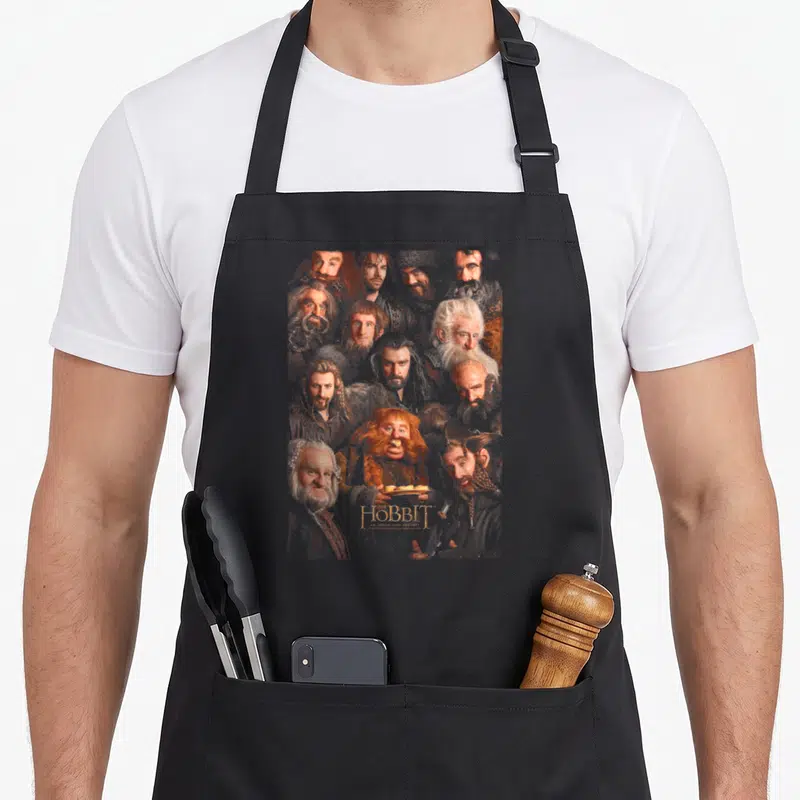 Tablier de cuisine noir avec une illustration regroupant les visages des nains et le logo doré du Hobbit en bas au centre.