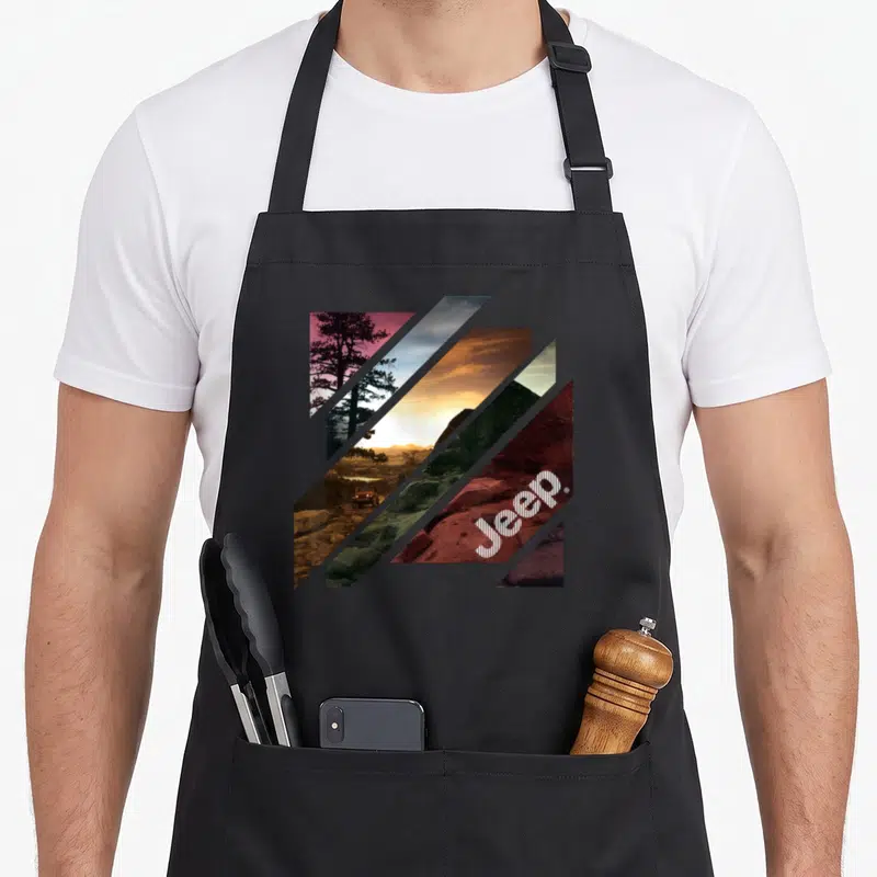 Tablier de cuisine noir avec motif géométrique diagonal montrant un véhicule sur terrain rocailleux et le logo Jeep stylisé.