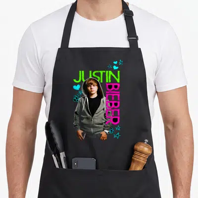 Tablier de cuisine noir avec Justin Bieber en haut gris, texte vert et rose, décor d'étoiles et cœurs bleus.