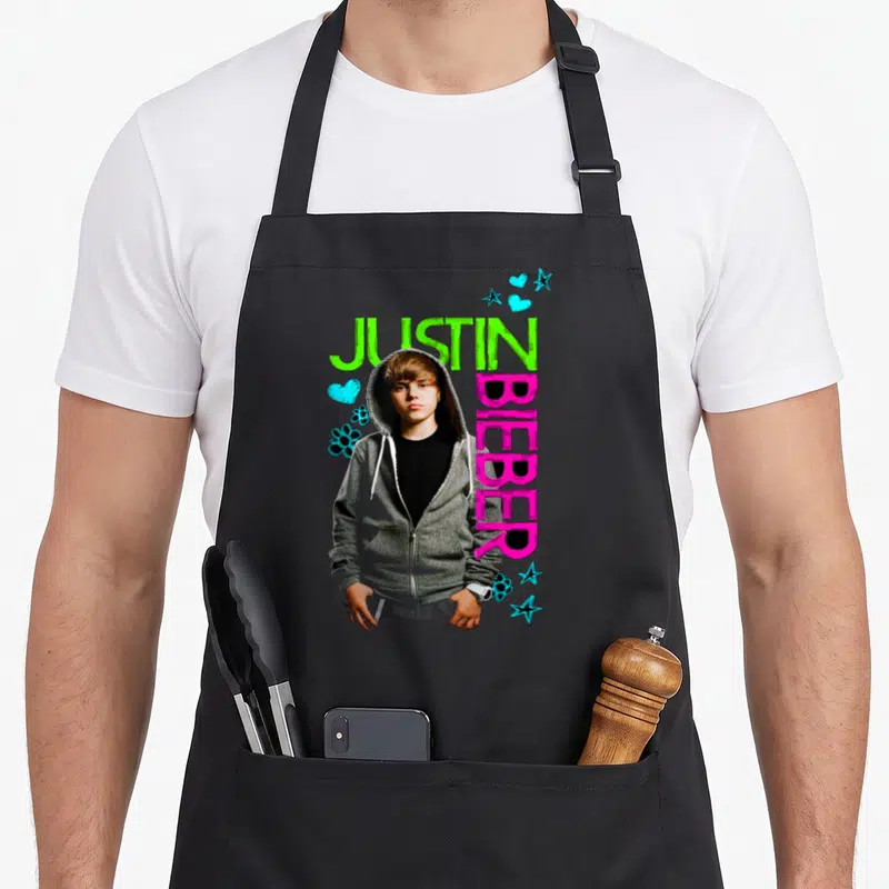 Tablier de cuisine noir avec Justin Bieber en haut gris, texte vert et rose, décor d'étoiles et cœurs bleus.