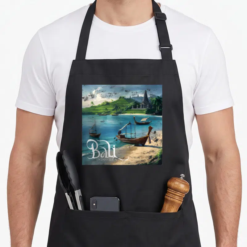 Tablier de cuisine noir avec une illustration colorée représentant des bateaux sur la plage de Bali, des rizières vertes et un temple indonésien sous un ciel nuageux.