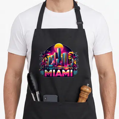 Tablier de cuisine noir avec illustration colorée du skyline de Miami, palmiers et texte MIAMI en lettres dégradées style synthwave.