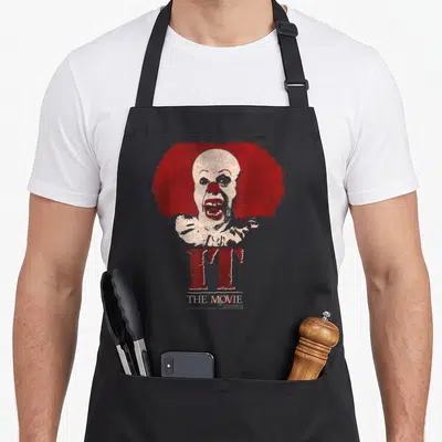 Tablier de cuisine noir avec illustration Pennywise Clown cheveux rouges et texte IT THE MOVIE en rouge et blanc.