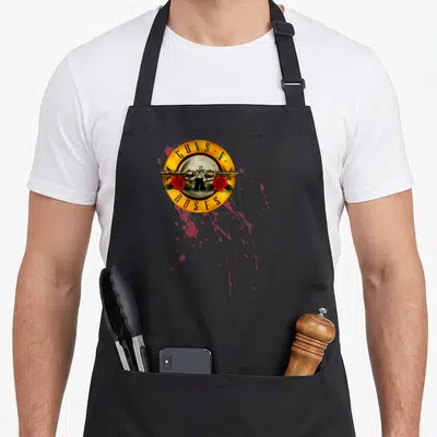 Tablier de cuisine noir avec logo circulaire jaune, deux revolvers, roses rouges et éclaboussages roses sur fond uni.