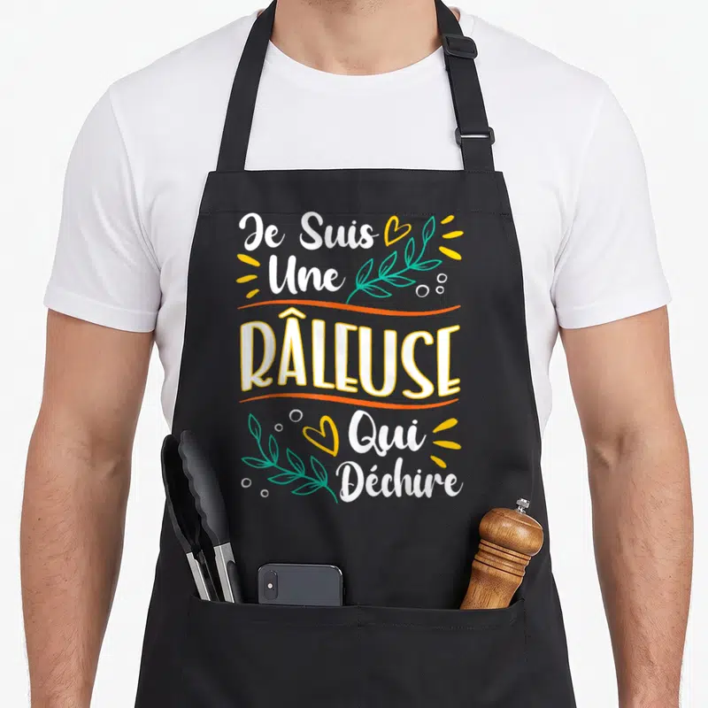 Tablier de cuisine noir avec l'inscription Je Suis Une Râleuse Qui Déchire en blanc et jaune entouré de feuilles vertes.