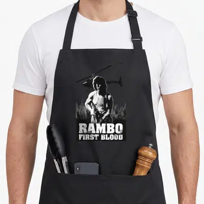 Tablier de cuisine noir avec illustration Rambo First Blood, hélicoptère et forêt en arrière-plan.