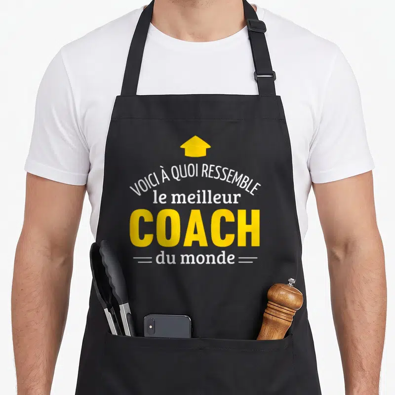 Tablier de cuisine noir avec inscription voici à quoi ressemble le meilleur coach du monde et une flèche jaune pointant vers le haut.