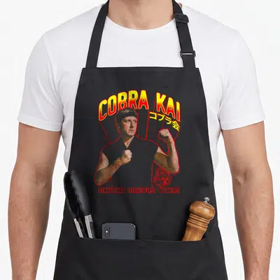 Tablier de cuisine noir avec illustration de Johnny Lawrence et texte Cobra Kai en jaune et rouge.