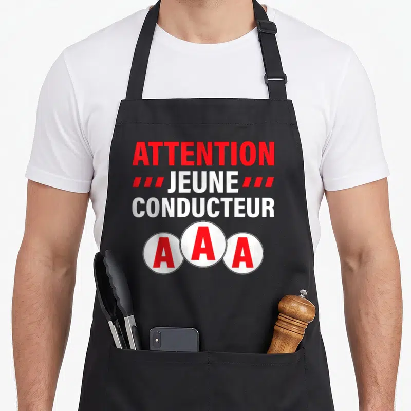 Tablier de cuisine noir avec inscription Attention Jeune Conducteur et trois badges AAA rouges et blancs.