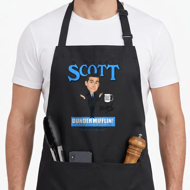 Tablier de cuisine noir avec caricature de Michael Scott en costume tenant une tasse et le logo Dunder Mifflin en bleu.