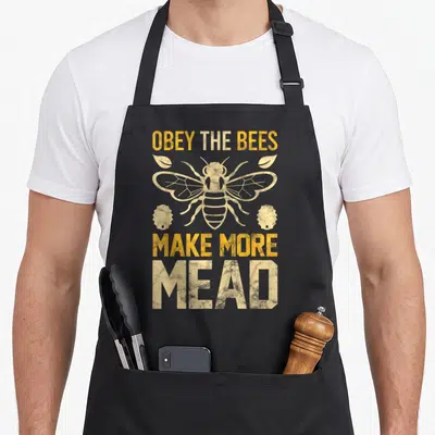 Tablier de cuisine noir avec une illustration d'abeille et le texte Obey the Bees Make More Mead en typographie jaune usée.