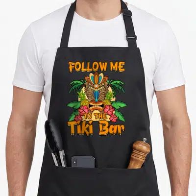 Tablier de cuisine noir avec un grand motif tiki tropicalFollow Me to The Tiki Bar entouré de fleurs et d'ananas.