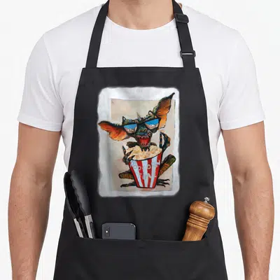 Tablier de cuisine noir avec illustration d'un monstre aux grandes oreilles mangeant du popcorn dans un pot rayé rouge et blanc.