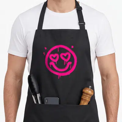 Tablier de cuisine noir avec grand smiley graffiti rose vif aux yeux en forme de cœur et effet de peinture coulante.