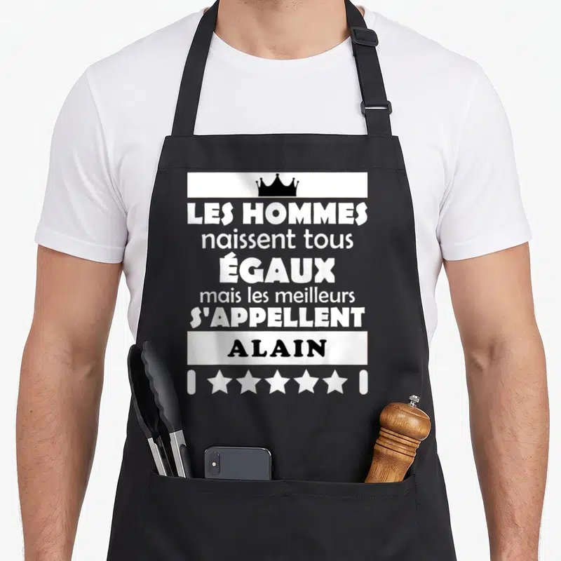 Tablier de cuisine noir avec une citation humoristique sur le prénom Alain et une couronne au-dessus.