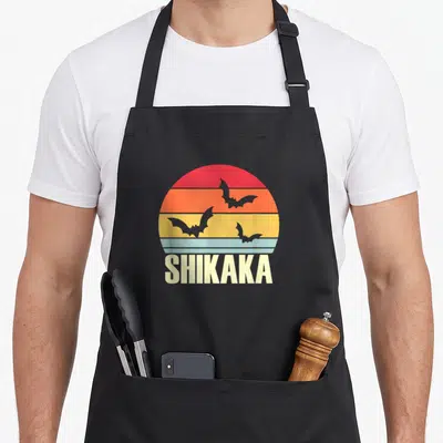 Tablier de cuisine noir avec un soleil rétro aux couleurs chaudes et trois chauves-souris volant devant le texte SHIKAKA en bas.