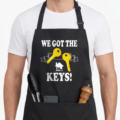 Tablier de cuisine noir avec deux clés jaunes croisées au-dessus d'une silhouette de maison blanche et du texte WE GOT THE KEYS en blanc.