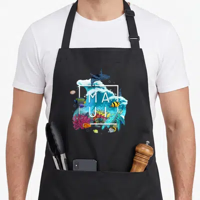 Tablier de cuisine noir avec illustration marine colorée incluant baleines, tortue et poissons autour du texte MAUI dans un cadre blanc.