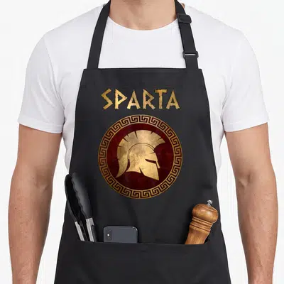 Tablier de cuisine noir avec inscription SPARTA et illustration de casque corinthien doré sur fond rouge circulaire.