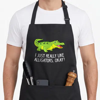 Tablier de cuisine noir avec un crocodile vert souriant et le texte I Just Really Like Alligators Okay imprimé en blanc.