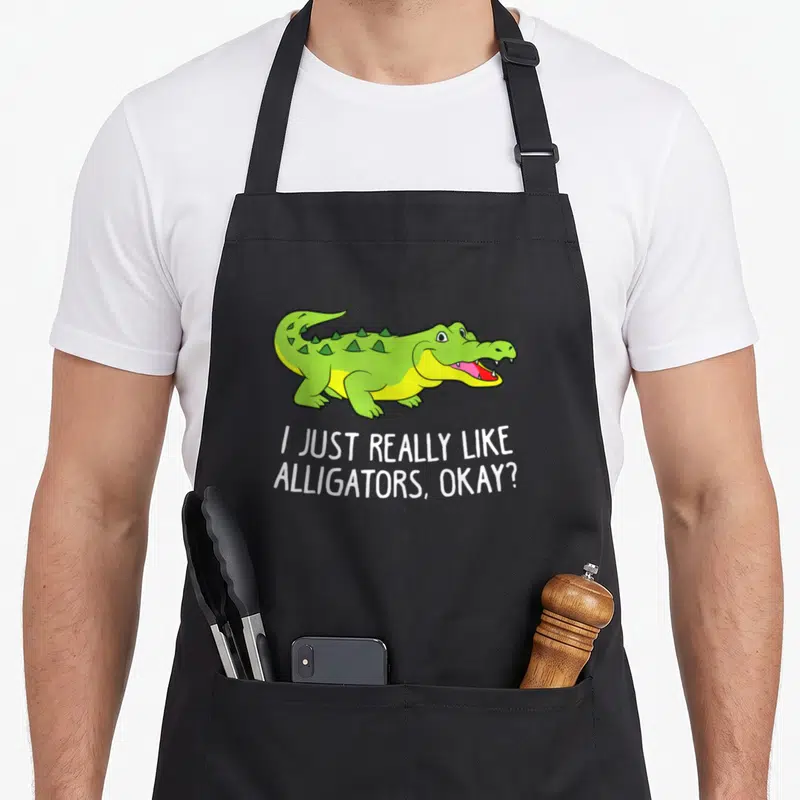 Tablier de cuisine noir avec un crocodile vert souriant et le texte I Just Really Like Alligators Okay imprimé en blanc.