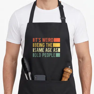 Tablier de cuisine noir avec le texte « It's Weird Being The Same Age As Old Folks » en lettres colorées style vintage.