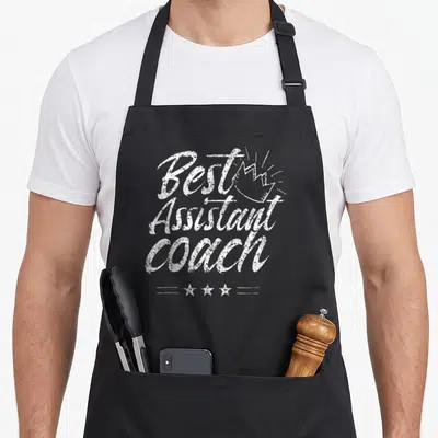 Tablier de cuisine noir avec inscription Best Assistant Coach et couronne dessinée en blanc.