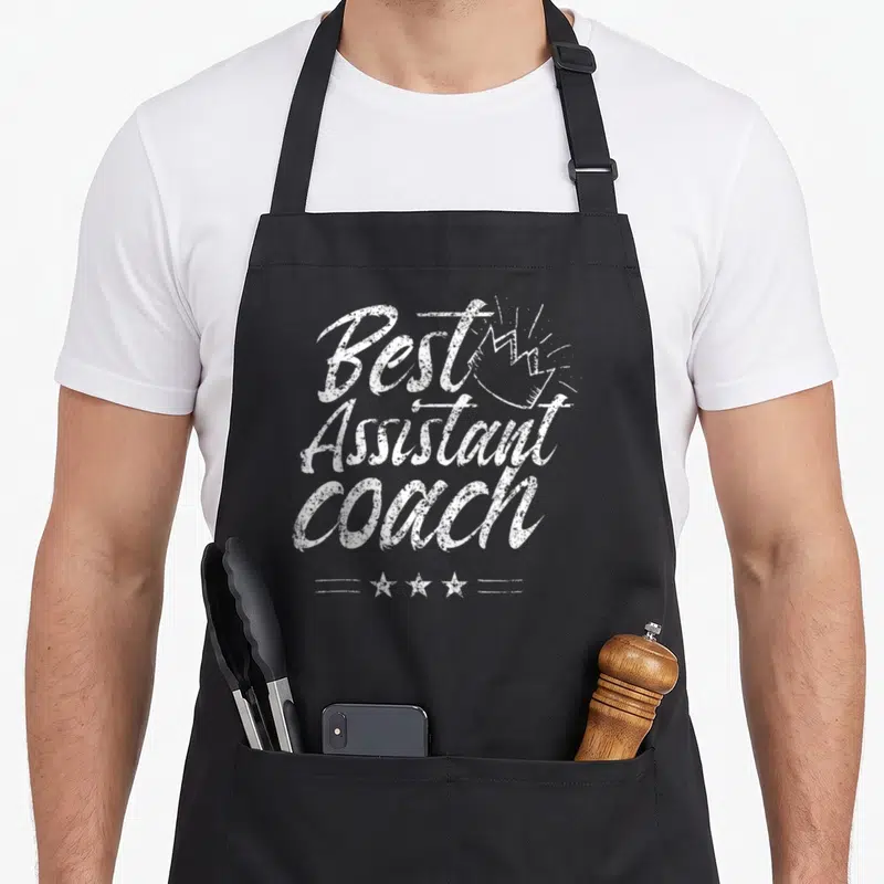 Tablier de cuisine noir avec inscription Best Assistant Coach et couronne dessinée en blanc.