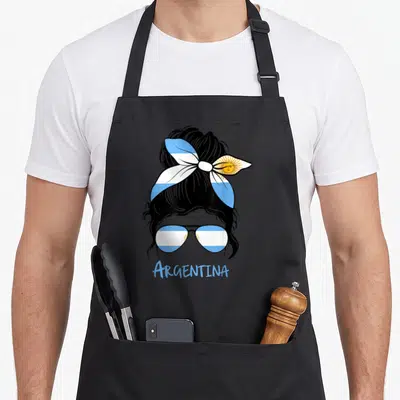Tablier de cuisine noir avec illustration de femme au chignon, ruban tricolore et lunettes aux motifs argentins.