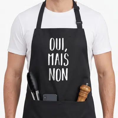 Tablier de cuisine noir avec l'inscription blanche OUI, MAIS NON en lettres capitales manuscrites.